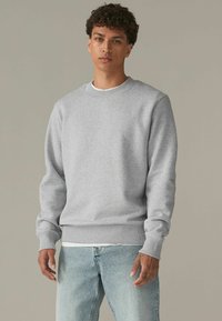 Grijze sweatshirt met ronde hals, lange mouwen, geribbelde manchetten en zoom, gedragen over een wit shirt, gecombineerd met lichtblauwe jeans.