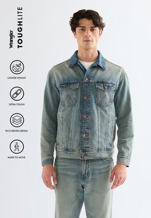 Uomo che indossa giacca di jeans chiara e jeans, in piedi contro uno sfondo semplice, con icone che evidenziano leggerezza, robustezza e flessibilità.