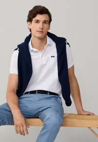 Jeune homme assis sur un banc en bois, portant un polo blanc, un pantalon bleu clair et un pull marine drapé sur les épaules.
