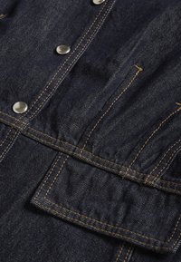 Tela de mezclilla azul oscuro con textura vertical prominente, acentuada con costuras naranja y botones a presión plateados. Incluye detalle de bolsillo delantero.