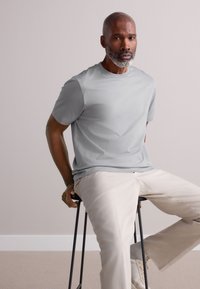 Világosszürke rövid ujjú póló kerek nyakkivágással, világos bézs nadrággal párosítva, fekete zsámolyon ülő személy által viselve.