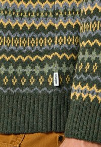 Sweter z wełny z dzianiny, ozdobiony geometrycznymi wzorami w kolorach zielonym, żółtym i szarym. Ściągacze przy mankietach i u dołu. Metka Wrangler na bocznej szwie.