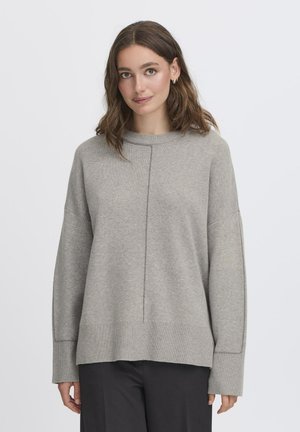 Donna con capelli castani fino alle spalle, che indossa orecchini a cerchio d'argento, un maglione grigio largo e pantaloni neri, in piedi contro uno sfondo chiaro.