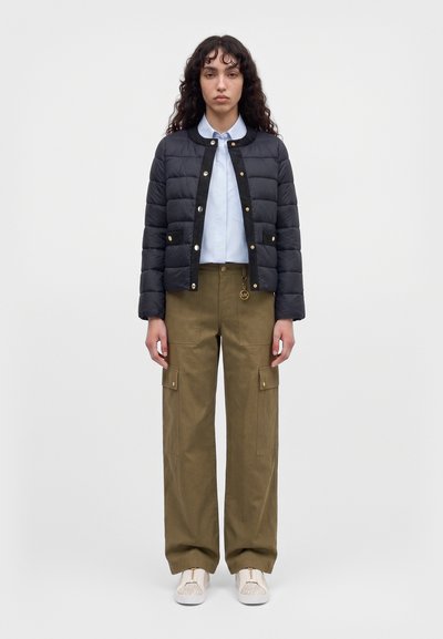 Une femme se tient face à l’objectif, vêtue d’une veste noire matelassée, d’une chemise blanche, d’un pantalon cargo olive et de baskets claires, sur un fond uni.