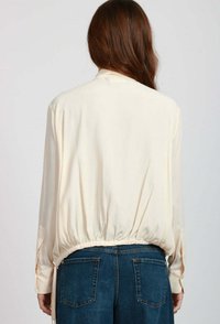 Blusa di crema con orlo arricciato, maniche lunghe e polsini con bottoni, abbinata a jeans in denim blu a vita alta, vista da dietro.