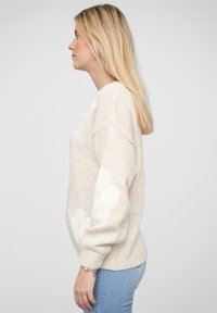 Hellbeige gestrickter Pullover mit lockerer Passform, der weiße Blumenmuster an den Ärmeln und einen runden Ausschnitt aufweist.