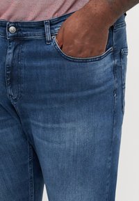 Mörkblå denimjeans med en slät textur. Har en framficka, subtila blekningar och en metallknappstängning.