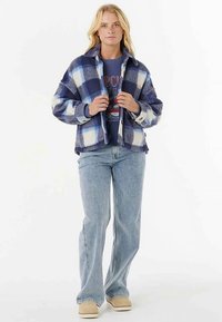 Marineblå og hvidternet fleecejakke med krave, båret over en blå grafisk t-shirt, kombineret med lyseblå flared jeans og beige sko.