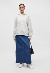 Sweatshirt cinza com um bolso frontal, combinado com uma saia longa de ganga, ténis brancos e uma bolsa preta. Modelo está de pé contra um fundo neutro.