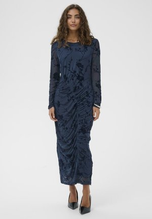 Frau, die ein langärmliges, navyblaues Kleid mit floralen Mustern und Raffung trägt, kombiniert mit schwarzen High Heels, steht vor einem weißen Hintergrund.