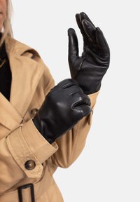 Schwarze Lederhandschuhe mit glatter Textur, einem eng anliegenden Design und drei sichtbaren Nähten auf der Rückseite. Getragen von einer Person in einem hellbraunen Mantel.