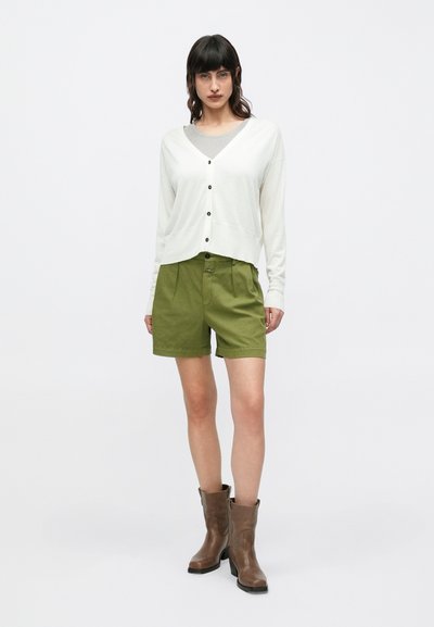 Γυναίκα που στέκεται φορώντας λευκό ζακέτα με κουμπιά πάνω από γκρι τοπ, χακί σορτς και καφέ μπότες μέχρι τη μέση της γάμπας, σε απλό φόντο.
