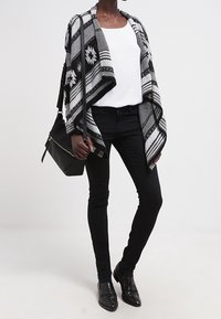 Černobílý pončo s vzorem a třásněmi, doplněný bílým topem a černými džínami. Černá kabelka a boty dokreslují outfit.