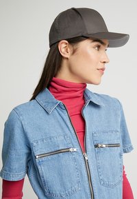 Tuta in denim a maniche corte, di colore azzurro chiaro, con due tasche con zip. Abbinata a un cappellino da baseball grigio scuro.