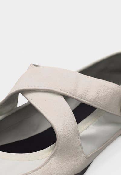 Μπεζ σουέντ σανδάλι με φαρδιές διασταυρούμενες λουρίδες και μαύρη μαξιλαρωμένη σόλα, φωτογραφημένο σε λευκό φόντο.