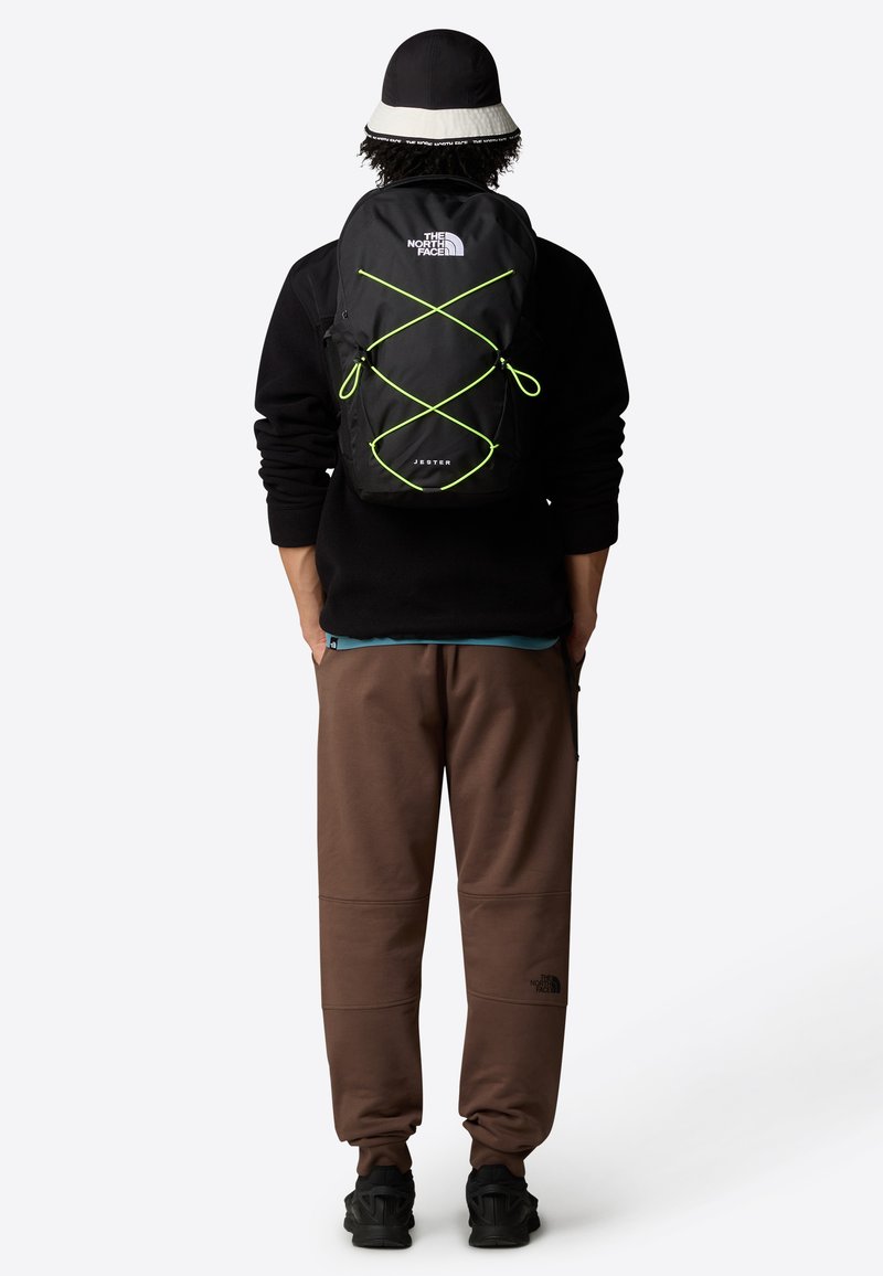 Musta reppu, jossa on neonvihreät yksityiskohdat, etupuolella bungee-köysimuotoilu ja logo. Yhdistetty ruskeisiin housuihin ja mustiin kenkiin.