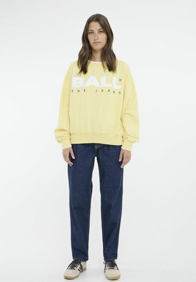 Gul sweatshirt med hvid "BALL" tekst, afslappet pasform, ribbede manchetter, parret med mørke denim jeans og afslappede sneakers. Minimalistisk design.