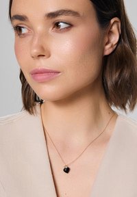 Złoty naszyjnik z czarnym sercem jako zawieszką, o gładkiej powierzchni i delikatnym łańcuszku, noszony na jasnej bluzce.