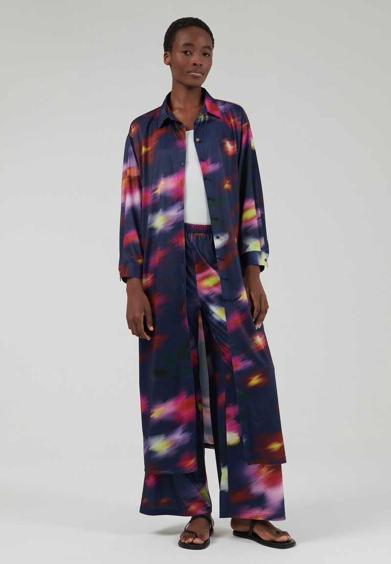 Lange, open voorkant blouse in donker marineblauw met een kleurrijk abstract patroon in roze, geel en groen. Gecombineerd met bijpassende wijde broek en sandalen.