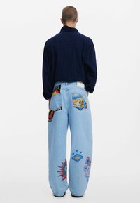 Hellblaue Jeans im weit geschnittenen Design, mit verschiedenen bunten Applikationen und Stickereien auf den hinteren Taschen. Kombinierbar mit einem navyblauen Texthemd.