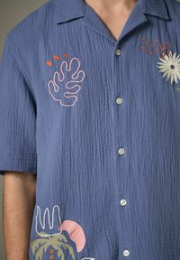 Kurzarmiges blaues Hemd aus strukturiertem Stoff, mit bunten gestickten Motiven von Blättern und Blumen sowie silbernen Knöpfen.