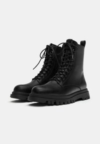 Fekete bőrből készült bokacsizma kör alakú orral, fűzős kialakítással, megerősített varrással és vastag gumi talppal, amely egy masszív futófelület mintázattal rendelkezik.