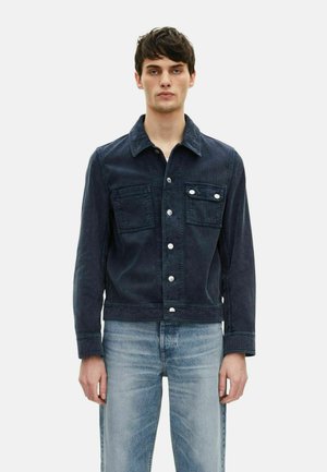 Veste en denim bleu foncé avec un col, deux poches avant et des boutons argentés. Associée à un jean bleu clair à coupe décontractée.