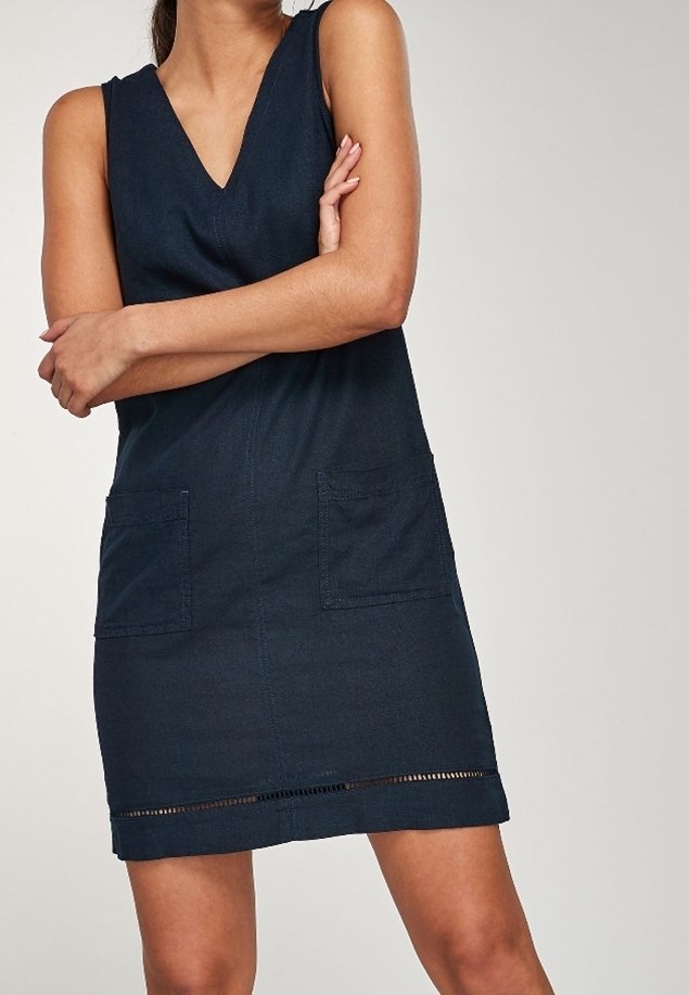 Femme portant une robe bleu foncé sans manches à encolure en V, avec des poches devant et un bandeau décoratif près de l’ourlet, debout les bras croisés.