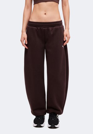 Personne portant un jogging large marron foncé, un top court et des chaussures de sport noires, debout devant un fond clair uni.