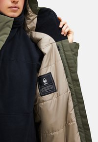 Gepolsterte Parka mit schwarzem Fleece und olivgrünen Akzenten. Verfügt über ein Innenetikett und eine Reißverschlusstasche. Reibungslose Texturen und gestepptes Design.