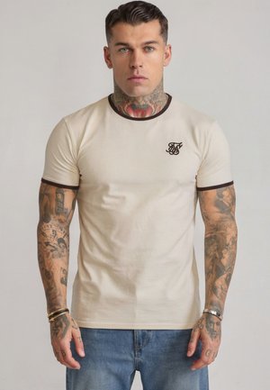Mann mit Hals- und Arm-Tätowierungen, der ein beiges T-Shirt mit dunklem Besatz und besticktem Logo trägt, steht vor einem schlichten hellen Hintergrund.