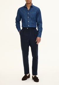 Dark blue denim button-up shirt with a chest pocket, paired with tailored dark trousers and brown suede shoes. Simple design and fit.