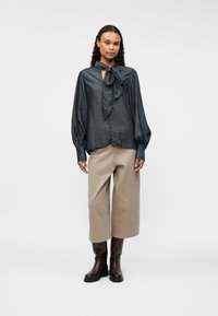 Tumši zila krekla ar kaklasaiti, garām pūkainām piedurknēm un gludu tekstūru. Apvienots ar pelēkiem platām biksēm un brūnām zābakiem.