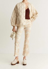 Chaqueta acolchada beige con patrón de diamantes, blusa de canalé color burdeos, pantalones vaqueros holgados de color claro con efecto de tie-dye y zuecos beige con detalles decorativos.