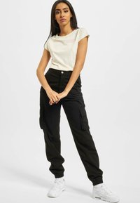 Tricou crem cu mânecă scurtă și un logo roz, asociat cu pantaloni cargo negri cu buzunare laterale și manșete elastice, purtat cu adidași albi.