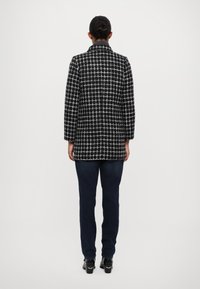 Abrigo a cuadros en blanco y negro con un diseño estructurado, dos bolsillos laterales y cuello. Portado con vaqueros azul oscuro y zapatos negros.