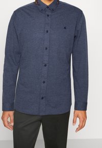 Marinblå långärmad skjorta med knappar i mjukt tyg, med krage och mörka knappar, som har en liten logotyp på bröstet.