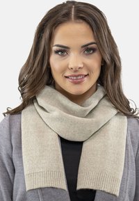 Krémová šála vyrobená z pleteného materiálu s měkkou texturou, s žebrovanými konci, elegantně draped kolem krku na šedém pozadí.