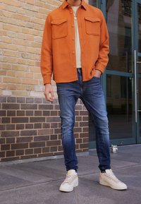 Chaqueta naranja de botones con dos bolsillos en el pecho, camiseta beige debajo, jeans ajustados azules y zapatillas beige claras. Fondo de pared de ladrillos.