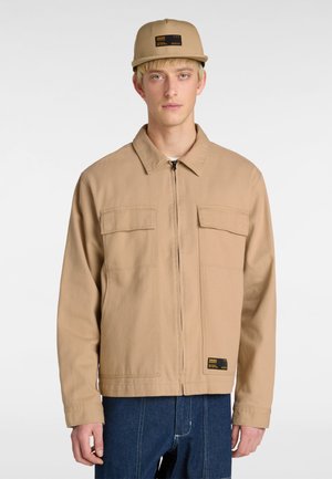 Giacca in tela color sabbia con zip frontale, due tasche sul petto e patch del marchio nero. Indossata con un berretto abbinato e jeans blu.