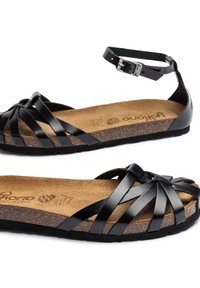 Sandalias de cuero negro con tiras entrecruzadas y hebilla en el tobillo. Plantilla de corcho, suela de goma negra texturizada y diseño plano para mayor comodidad.