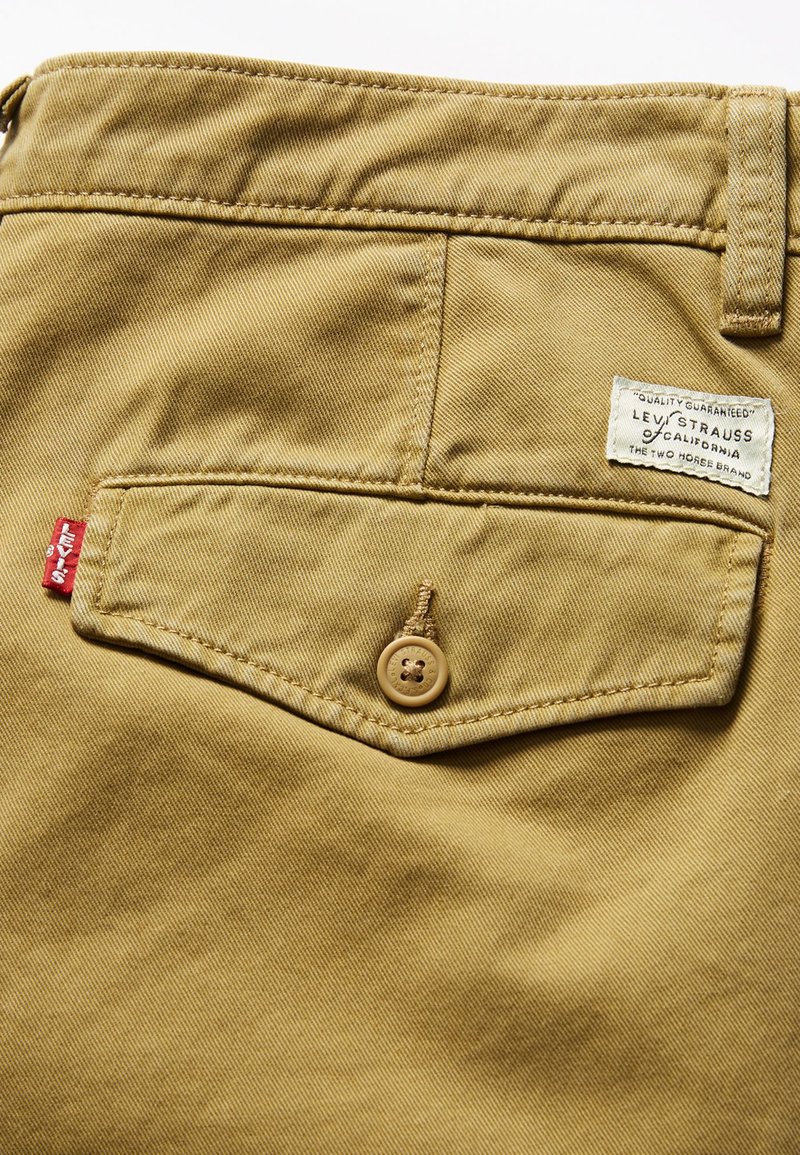 Tela de algodón beige con un bolsillo trasero que presenta un botón marrón claro. Incluye una etiqueta con "Levi Strauss & Co." y una etiqueta roja de marca.