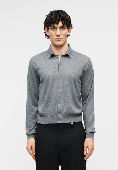 Jeune homme aux cheveux bouclés portant une chemise grise à manches longues boutonnée et un pantalon noir, debout devant un fond clair uni.