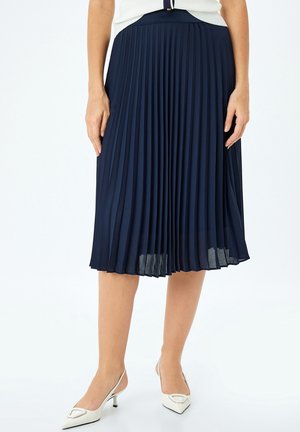 Marineblauwe geplooide rok met een gladde textuur, knielang, voorzien van een aansluitende tailleband en subtiele glans, gecombineerd met witte slingback hakken.