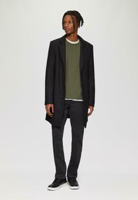 Schwarzer Mantel mit Revers, grüner Rippstrickpullover, schwarze Jeans und schwarze Sneaker mit weißen Sohlen. Lässiges, schichtartiges Outfit.