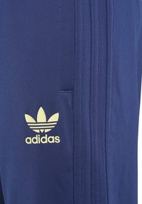 Jūras spēku zilas sporta bikses, kas izgatavotas no teksturēta auduma, ar trim vertikālām svītrām, ar rāvējslēdzēju kabatu un dzelteno adidas logo pa kreisi pie augšstilba.