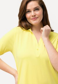 Geel polo shirt met korte mouwen, voorzien van een knoopsluiting en zachte, gladde stof. Model met halflang bruin haar.