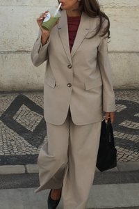 Traje beige a medida con pantalones de pierna ancha y chaqueta, que presenta dos botones. Complementado con una blusa burdeos y un bolso negro.