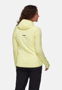 Helles gelbes Fleece-Hoodie mit taillierter Passform, Kapuze und schwarzem Logo auf dem oberen Rücken. Weiche Textur und lange Ärmel.