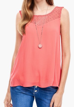 Femme portant un chemisier corail sans manches avec un détail en dentelle sur le yoke et un long collier avec pendentif rond, assorti à un jean bleu.